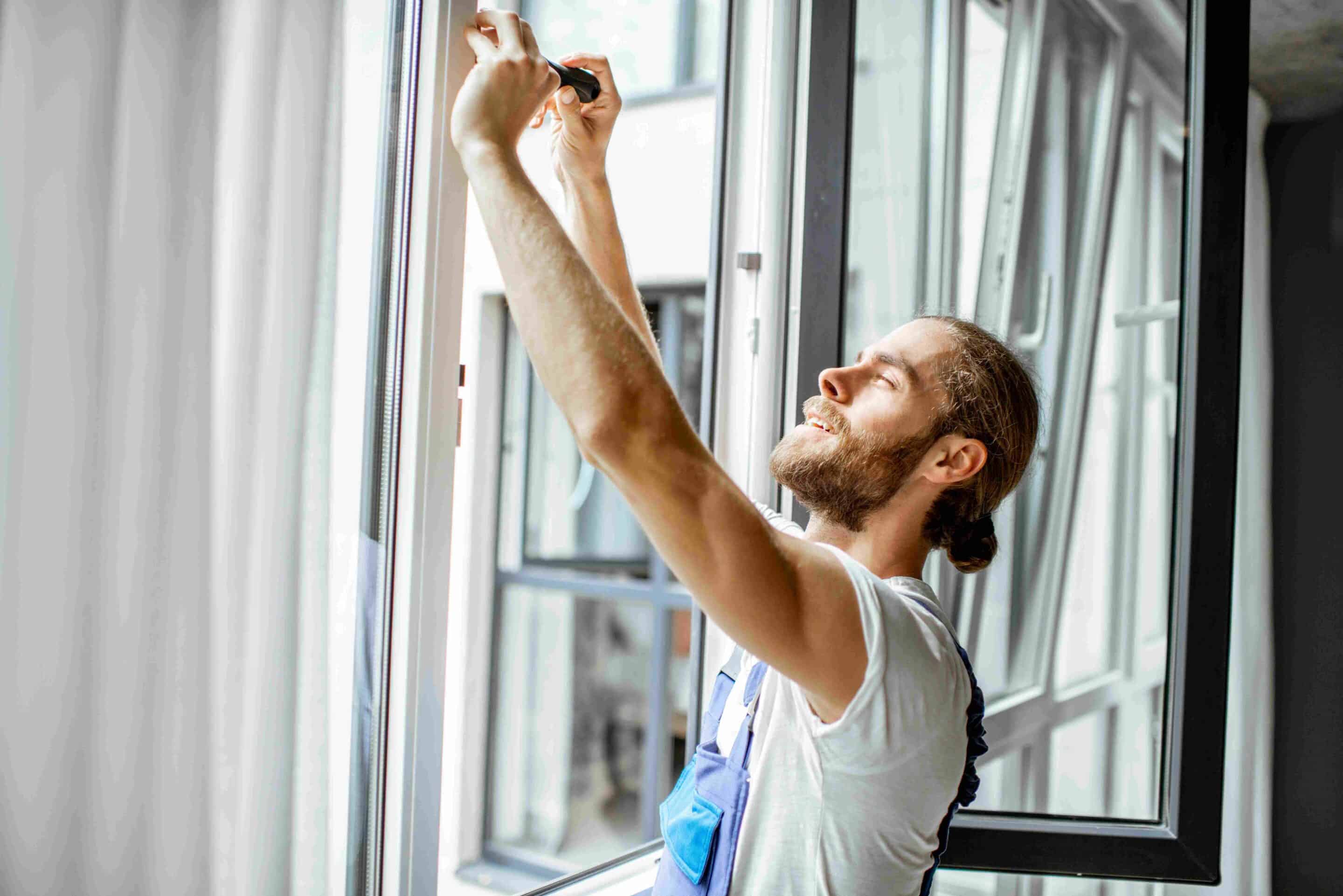 A smiling man in work overalls repairs or installs a window, using a tool while standing indoors near an open window with sunlight streaming in.