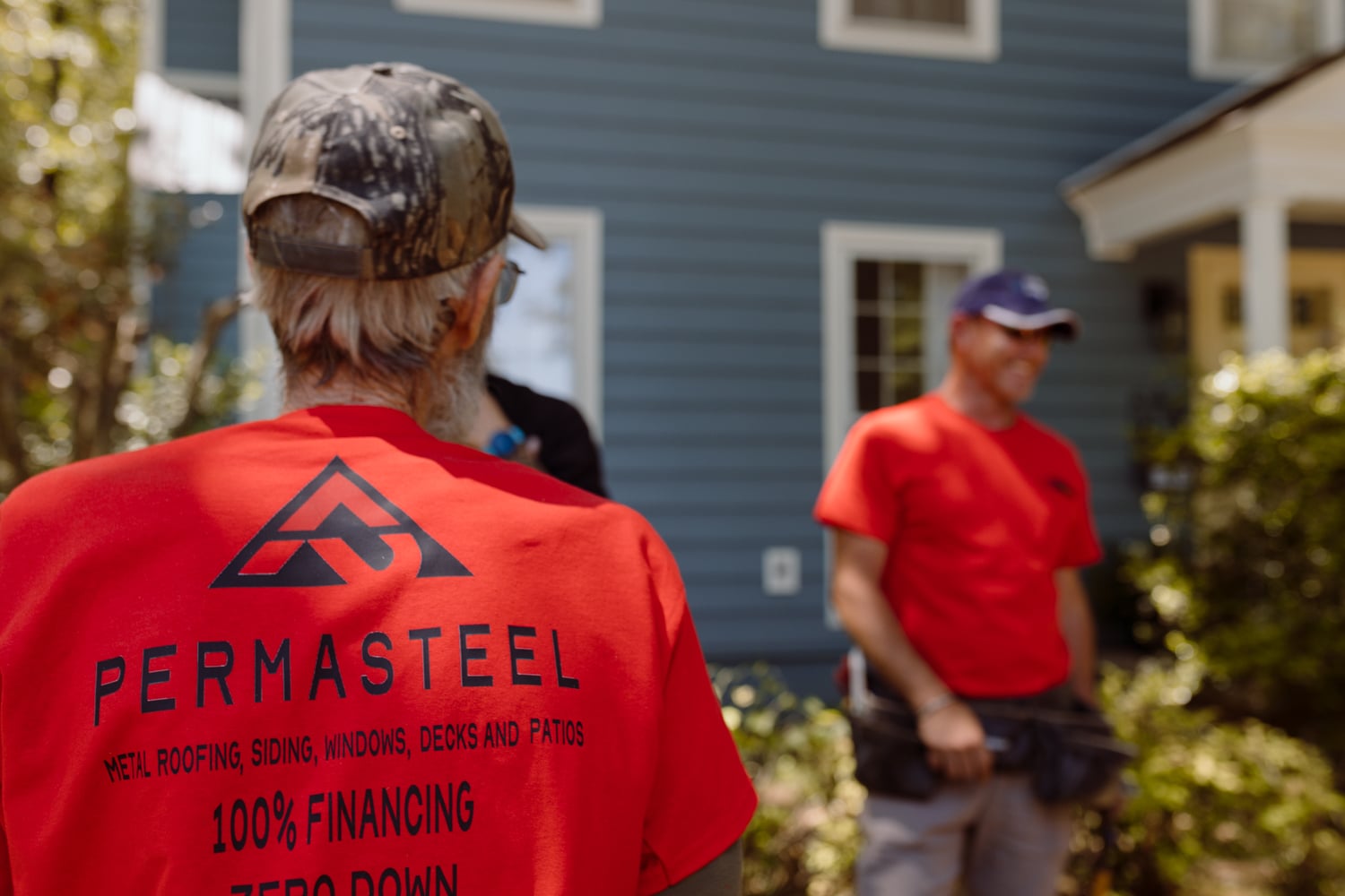 A man in a red PERMASTEEL shirt stands outdoors facing two other men near a blue house. The shirt advertises metal roofing and financing options. The scene appears casual and work-related.